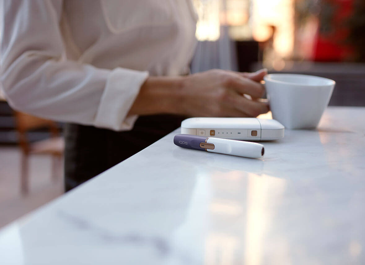 Mujer con IQOS 2.4 PLUS personalizado en violeta en un bar con un café