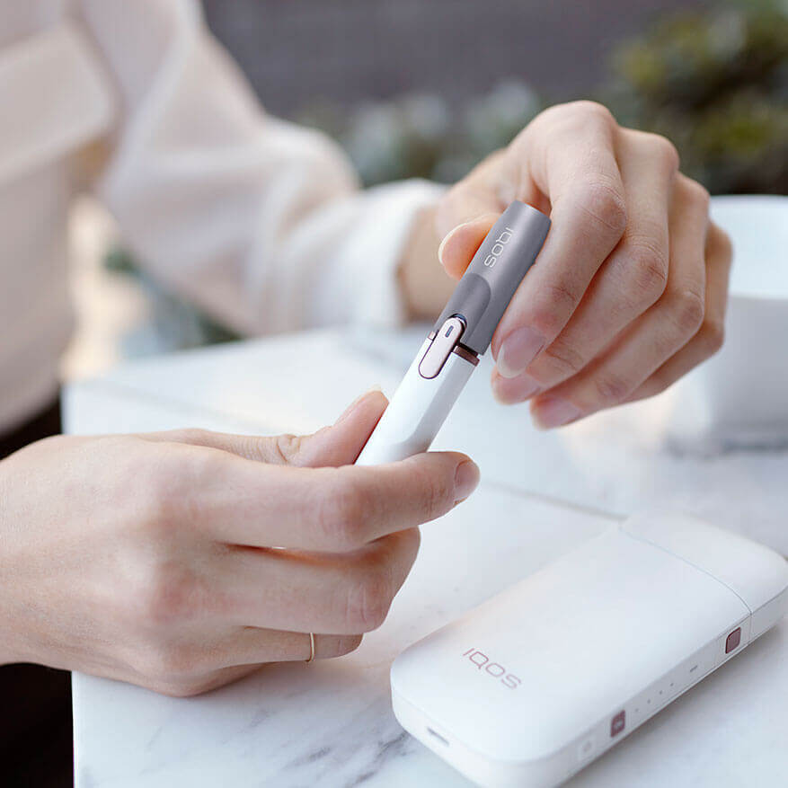 Mujer con IQOS 2.4 PLUS personalizado en gris en un bar con un café