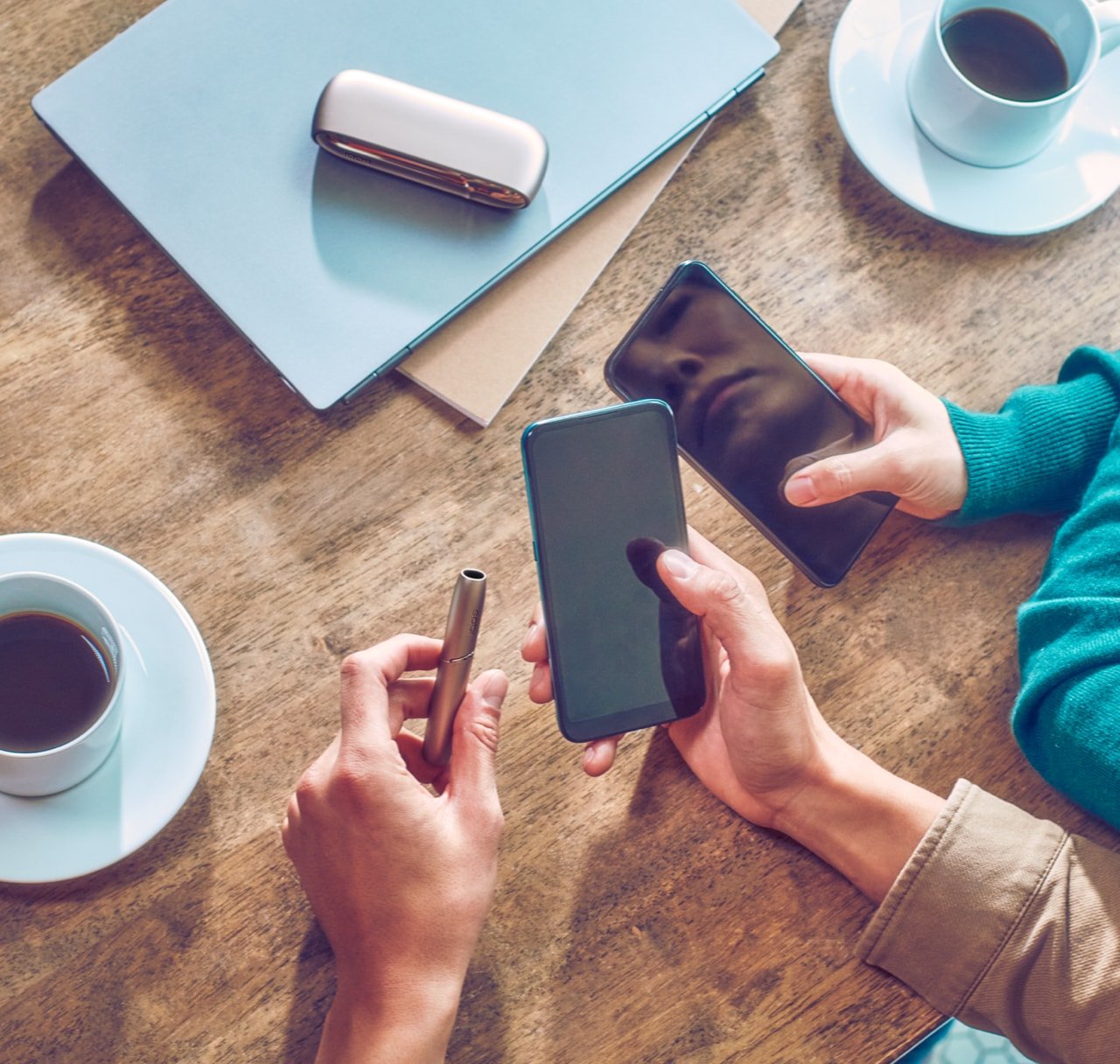 Two people holding mobile phones sit at a table; one also holds a jade blue IQOS ILUMA PRIME device.