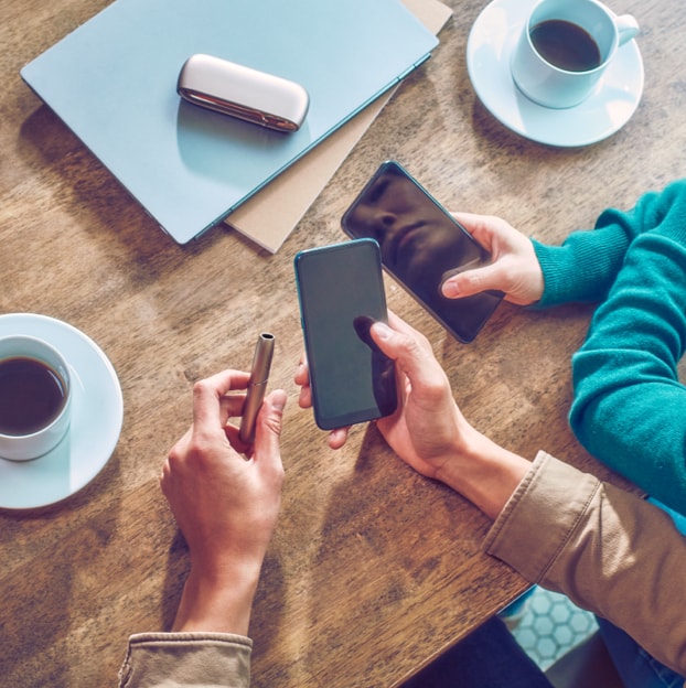 Two people holding mobile phones sit at a table; one also holds a jade blue IQOS ILUMA PRIME device.