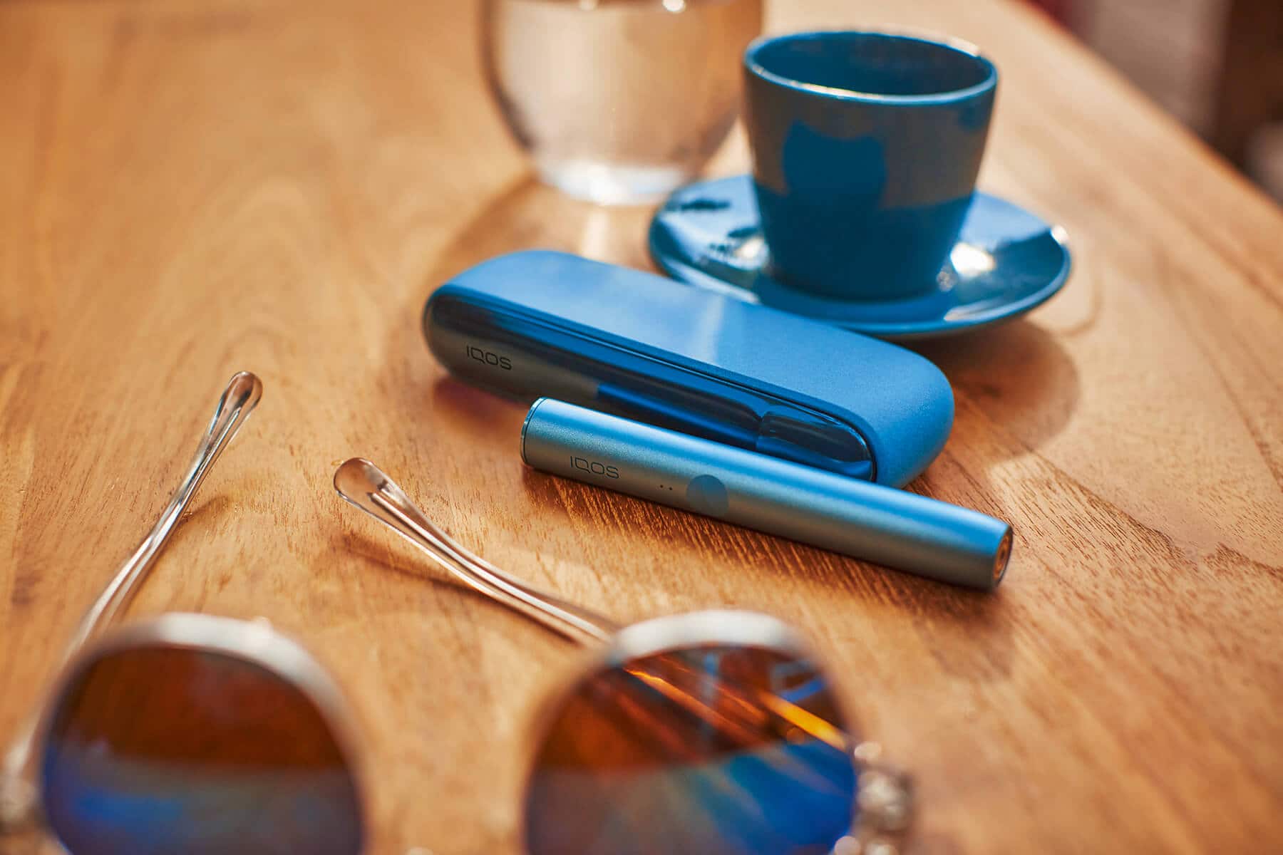 Dispositivo IQOS en mesa con gafas de sol y taza de cafe