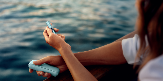 Mujer sujetando un IQOS ILUMA i con el mar de fondo