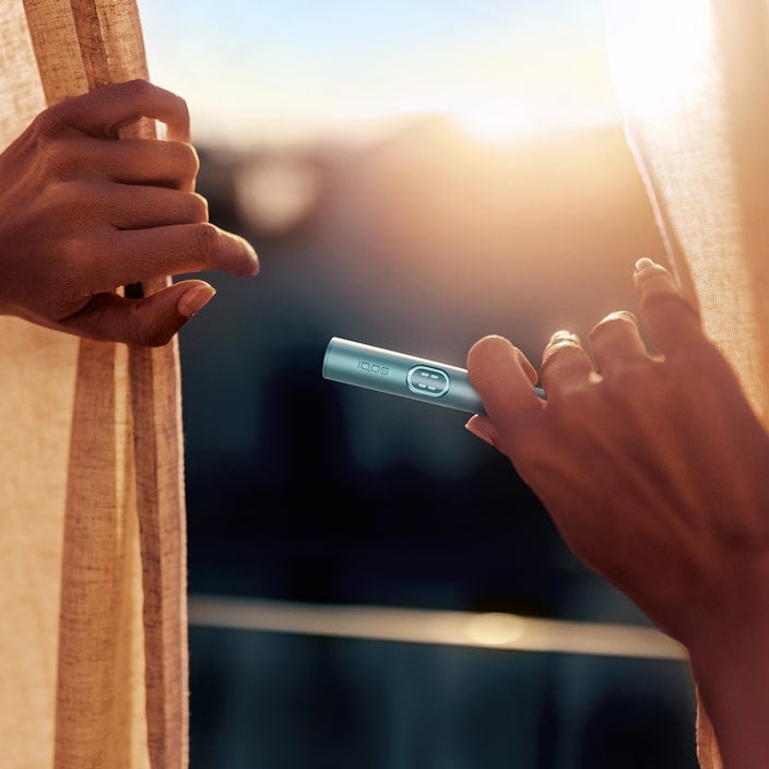 Dos manos sujetando unas cortinas con una puesta de sol de fondo y una de las manos sujetando el holder de IQOS ILUMA i PRIME en azul brisa