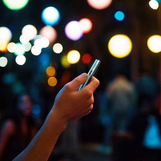 Mano sujetando Holder IQOS ILUMA i en Azul Brisa con luces de colores de fondo