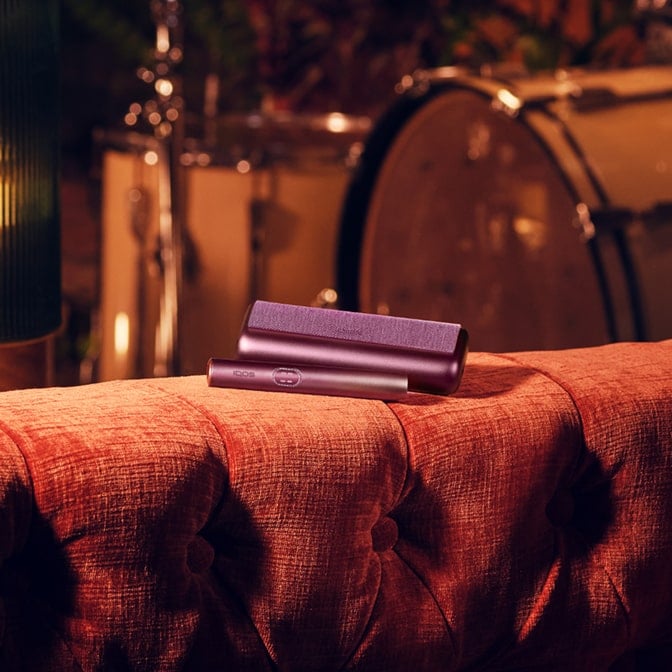 Dispositivo IQOS ILUMA i PRIME Rojo Granate encima de un sofa y una batería de fondo
