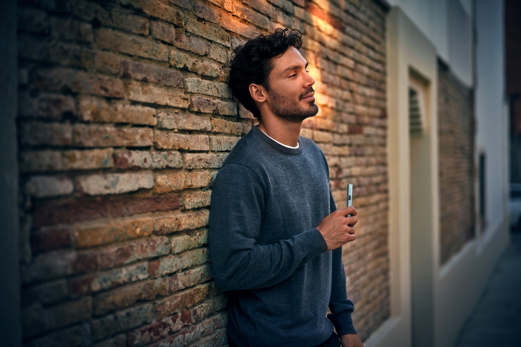 Hombre apoyado en una pared de ladrillos con holder de IQOS ILUMA i