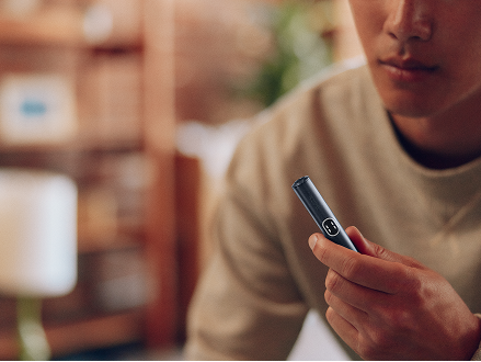 Hombre sujetando un dispositivo IQOS azul oscuro en interior, en primer plano, con fondo desenfocado y ambiente cálido.