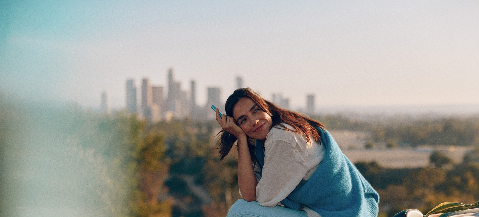 Mujer sentada al aire libre con vistas a la ciudad al fondo, sonriendo mientras sostiene un dispositivo IQOS azul claro en un ambiente relajado y luminoso.