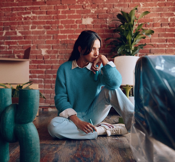Mujer sentada en el suelo de un loft con cajas de mudanza, sosteniendo un dispositivo IQOS azul claro en un ambiente interior acogedor con pared de ladrillo.