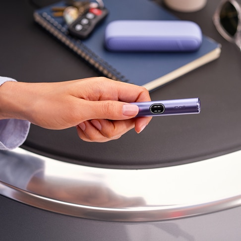 Mano sosteniendo un dispositivo electrónico morado sobre una mesa con libreta, gafas de sol y otros objetos personales al fondo.