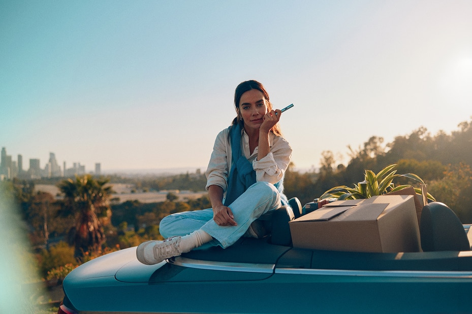 Mujer sentada sobre un coche descapotable con cajas de mudanza y plantas, sosteniendo un dispositivo IQOS azul claro, con vistas a la ciudad al fondo.