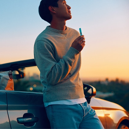 Hombre apoyado en un coche al atardecer, sosteniendo un dispositivo IQOS en la mano y mirando hacia el horizonte.