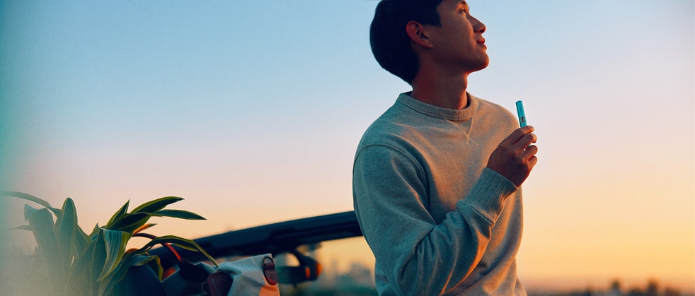 Persona de pie junto a un coche al atardecer, sosteniendo un dispositivo electrónico con paisaje desenfocado al fondo.