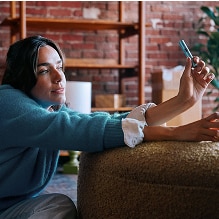 Mujer en interior observando un dispositivo IQOS azul claro que sostiene en la mano, con fondo de pared de ladrillo y ambiente acogedor