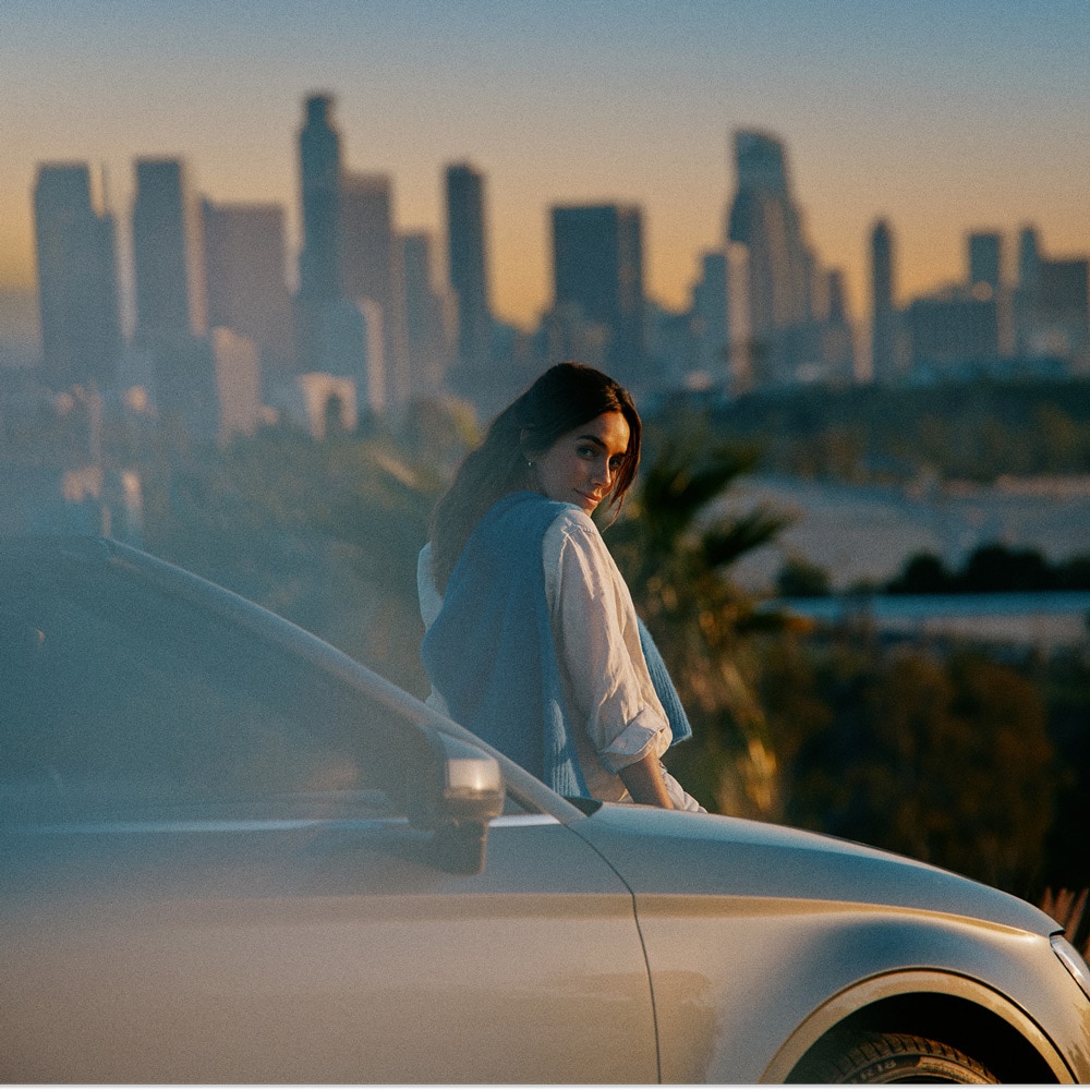 Mujer apoyada junto a un coche con el skyline de una ciudad al atardecer desenfocado al fondo.