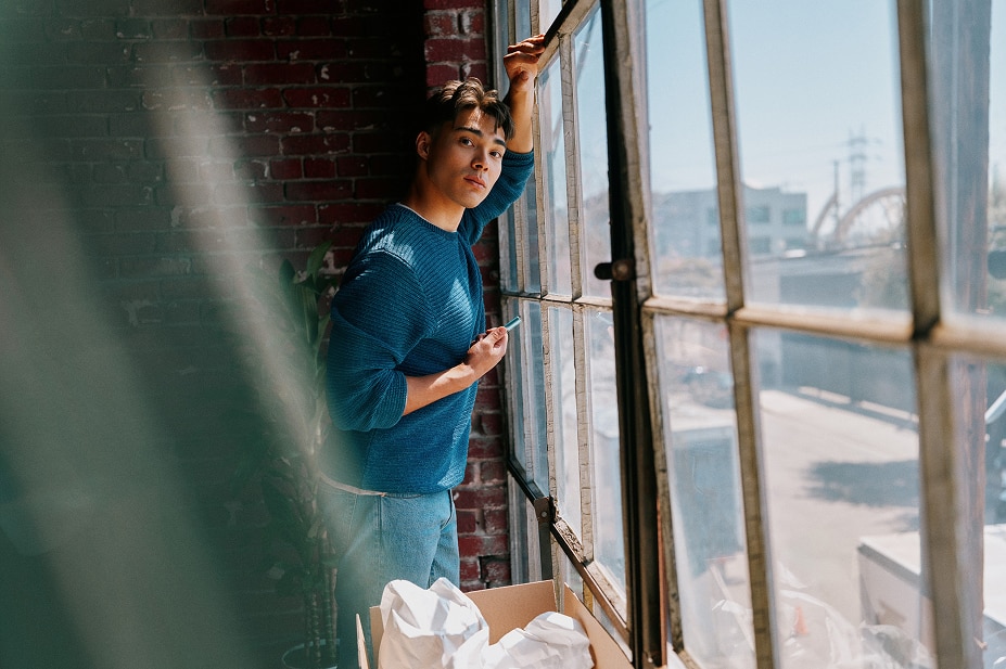 Hombre de pie junto a una ventana industrial, mirando a cámara mientras sostiene un dispositivo IQOS azul claro en un espacio interior con luz natural.