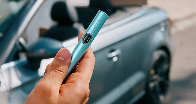 Persona sosteniendo un dispositivo IQOS turquesa junto a un coche descapotable en un entorno urbano.