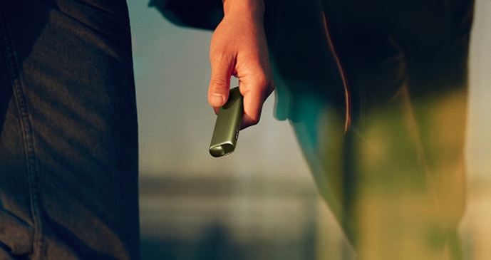 Primer plano de una mano sujetando un dispositivo IQOS verde en un entorno exterior con luz cálida
