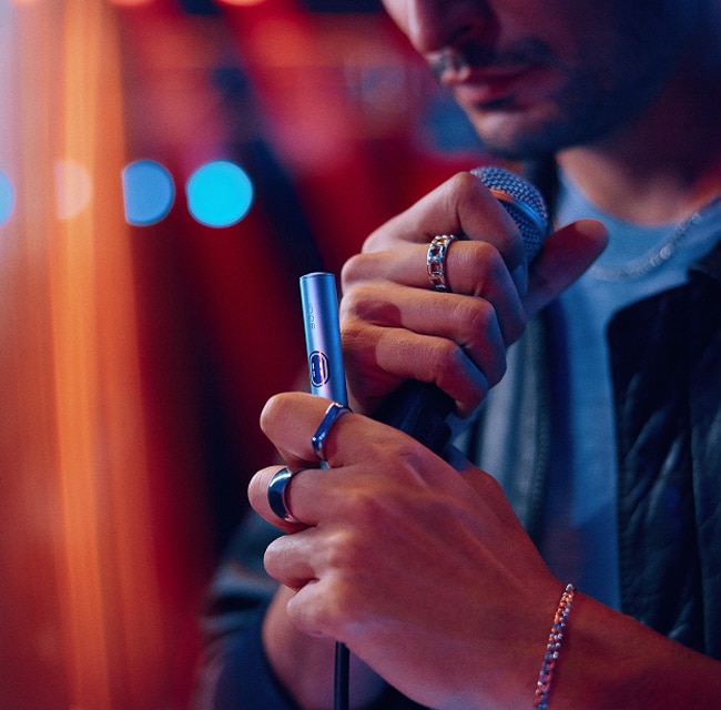 Primer plano de una persona sujetando un micrófono y un dispositivo IQOS plateado, con iluminación de neón en un ambiente nocturno.