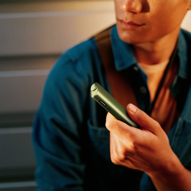 Hombre con camisa vaquera azul sosteniendo en la mano un dispositivo IQOS ILUMA i de color verde, en un ambiente con luz cálida y fondo de pared metálica.