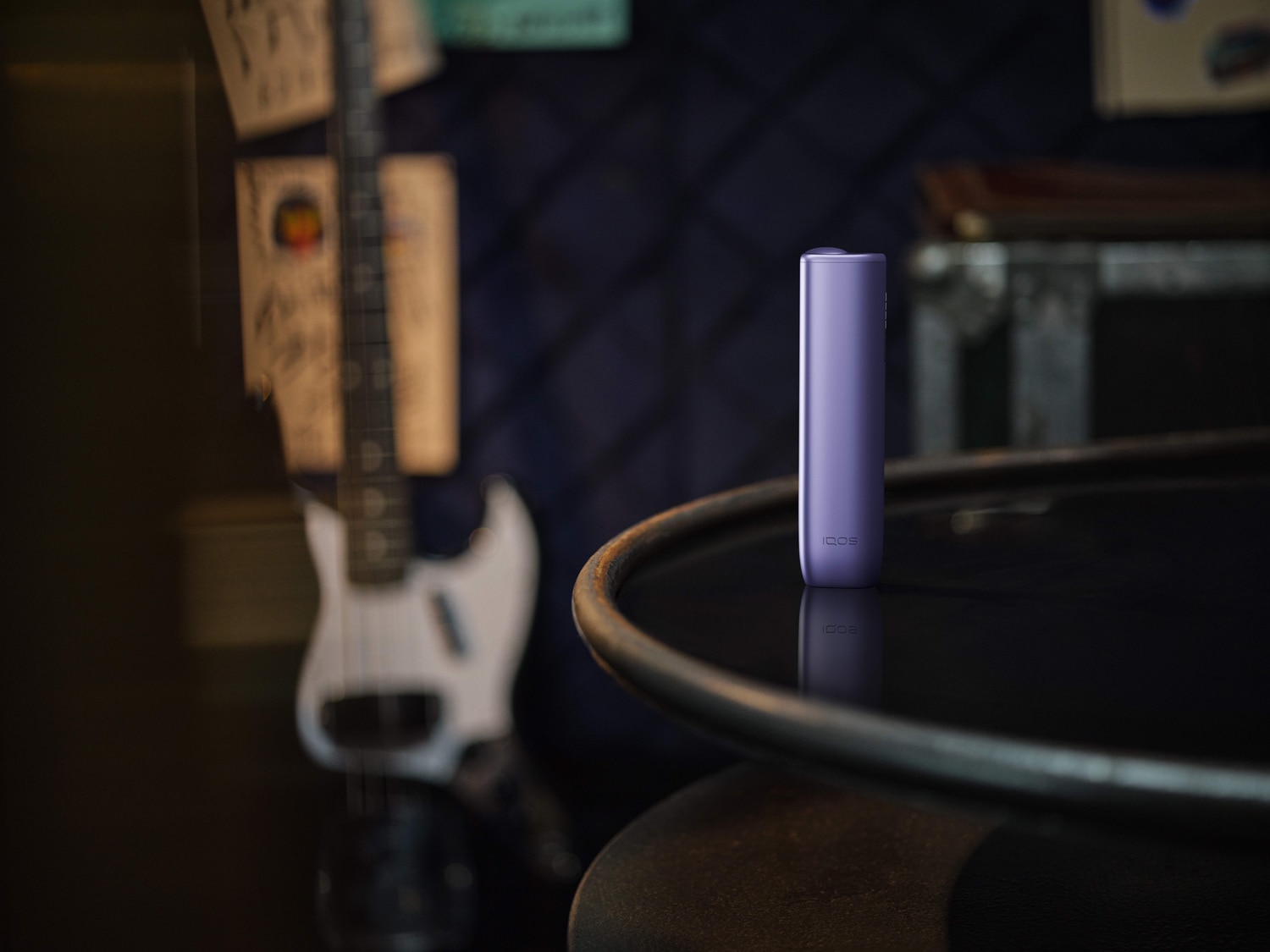 Dispositivo IQOS de color violeta colocado en posición vertical sobre una mesa redonda oscura, con una guitarra eléctrica blanca desenfocada al fondo en un ambiente tenue.