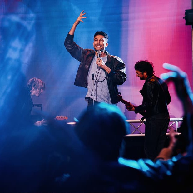 Cantante en el escenario con el brazo levantado y micrófono en mano, actuando ante el público. Al fondo, un guitarrista y un teclista. Iluminación dramática en tonos azul y rosa