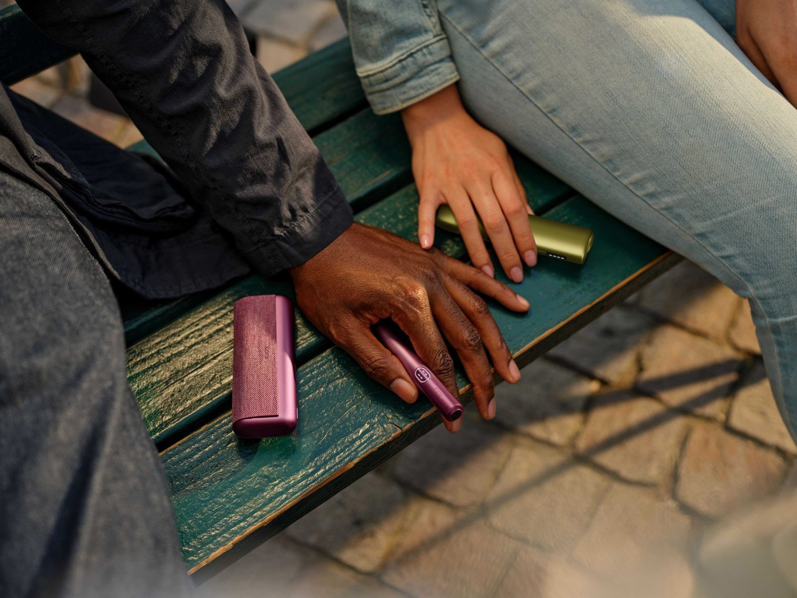 Two people sitting close together on a weathered green wooden bench, their hands resting beside IQOS devices in burgundy and olive-green finishes, with cobblestone paving in the background.