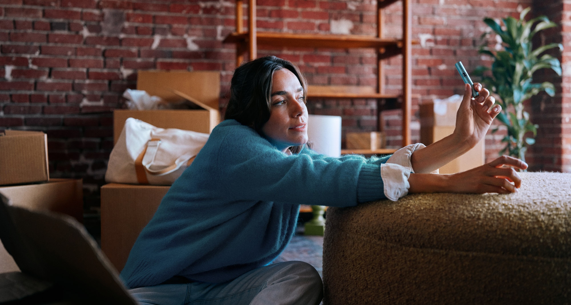 Woman in a teal sweater leaning on a pouffe, holding a turquoise IQOS device, surrounded by moving boxes in a brick-walled room