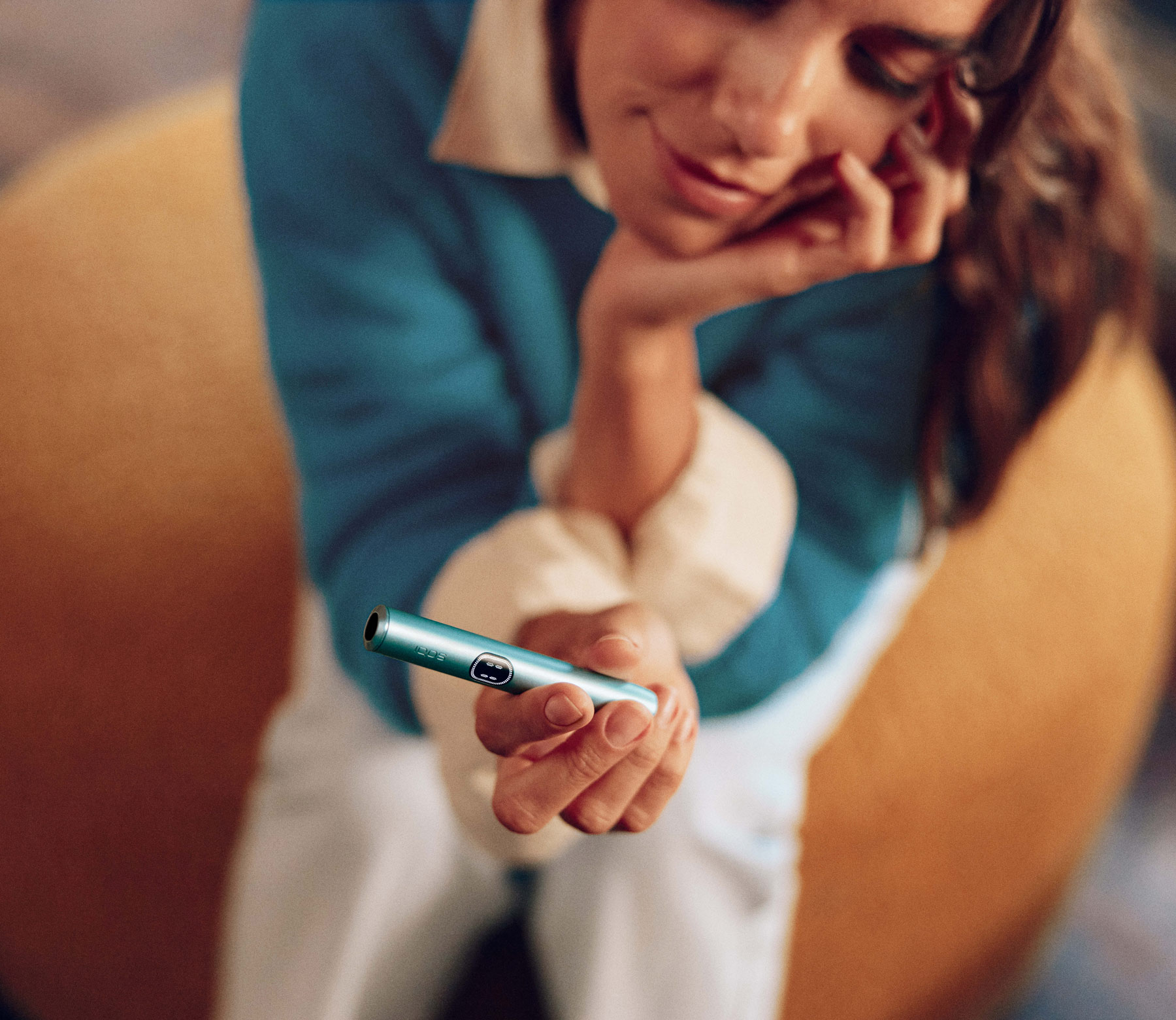 Smiling woman relaxing on a yellow beanbag, resting her chin on one hand while holding out a turquoise IQOS device in the other.