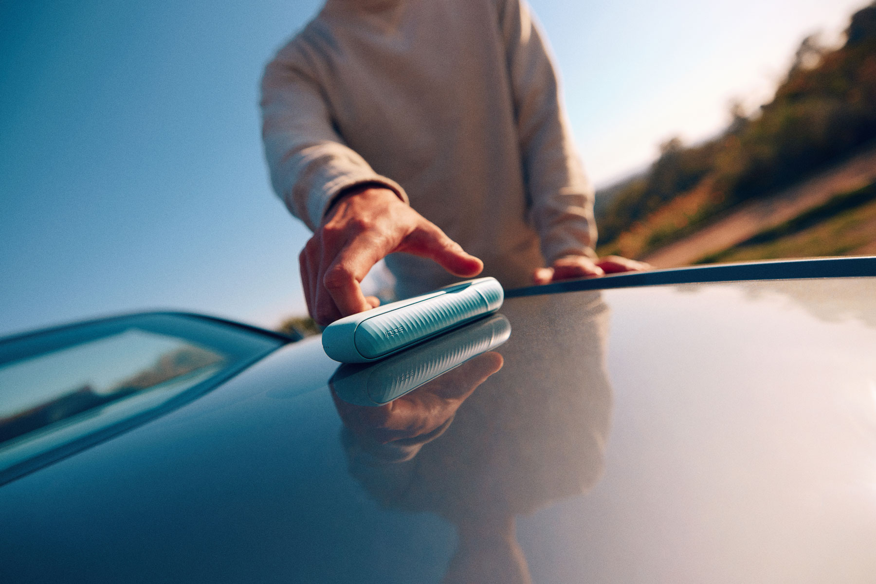 Usuario de IQOS cogiendo su dispositivo del capó de su coche en una escena de atardecer