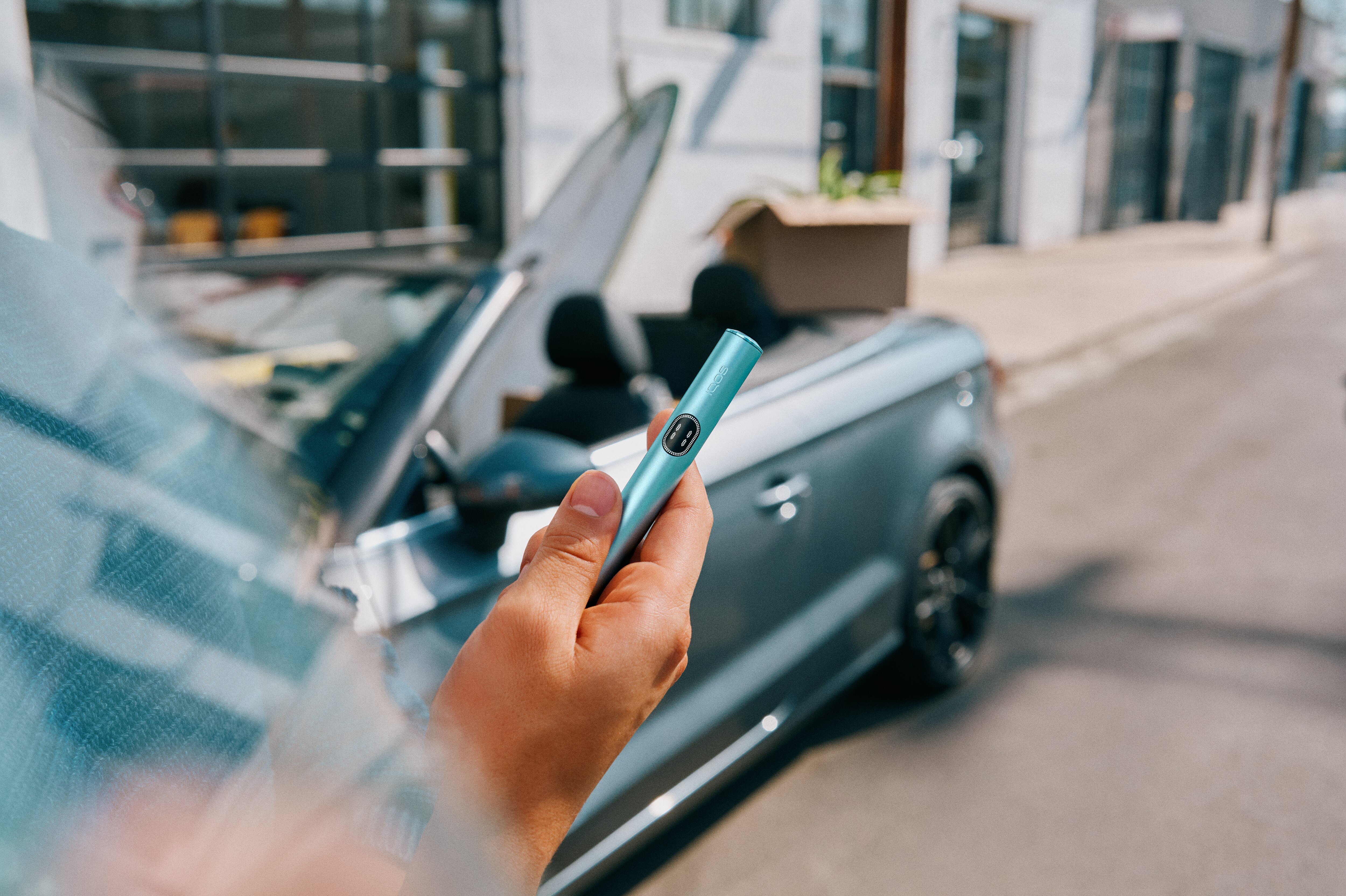 Mano sujetando un IQOS con un coche de fondo