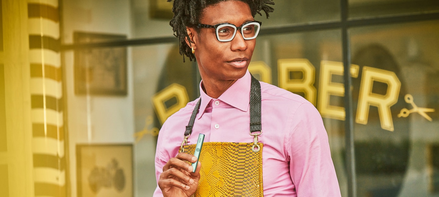 A person holding an IQOS device while standing in front of a barber shop.