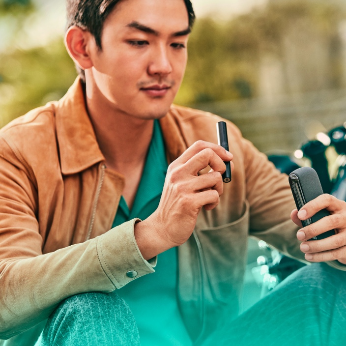A man holding an obsidian black IQOS ILUMA PRIME Holder while looking at his mobile phone.