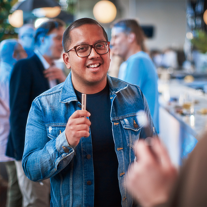 Smiling man dressed in blue and holding a silver IQOS device in his hand